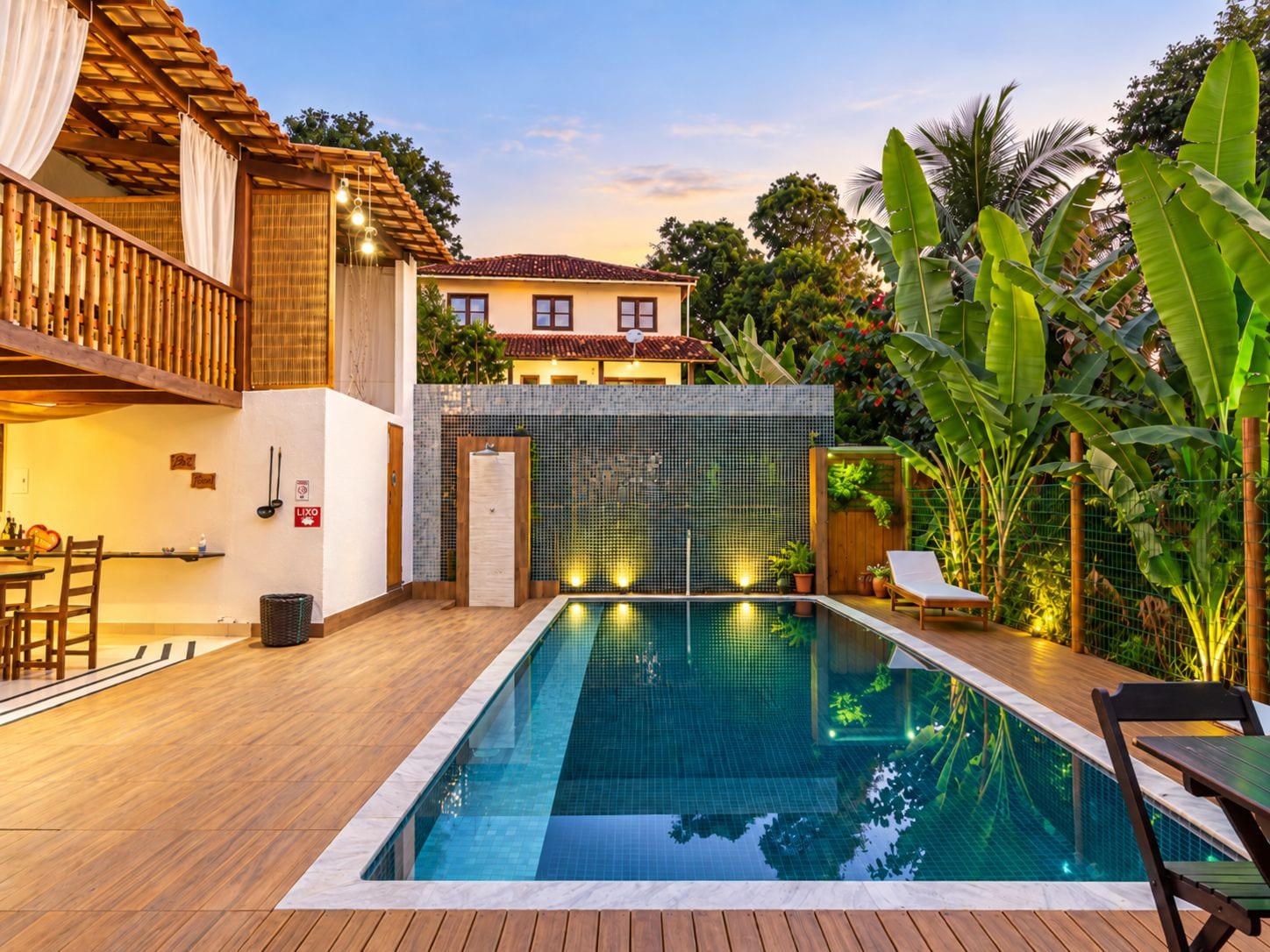 A swimming pool with a wooden deck, a covered seating area, and a view of a house in the background.