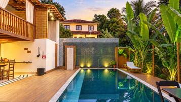 A swimming pool with a wooden deck, a covered seating area, and a view of a house in the background.