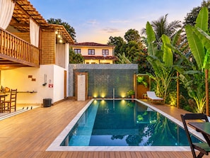 A swimming pool with a wooden deck, a covered seating area, and a view of a house in the background.