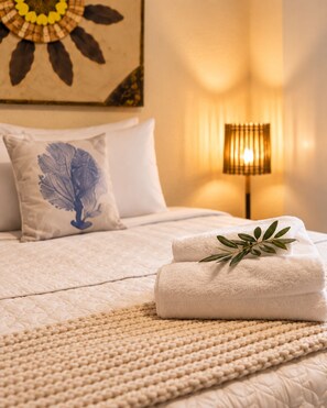 Una cama bien hecha con una cubierta beige texturizada, una almohada blanca con un diseño de hoja azul y una toalla blanca doblada con una hoja verde encima.