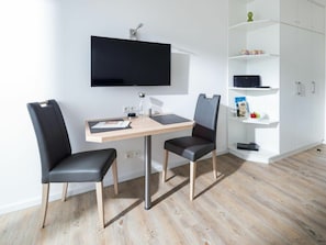 Ein modernes Zimmer mit einem Wand-Fernseher, einem Holztisch mit zwei Stühlen und einer weißen Regalkonstruktion mit unterschiedlichen Gegenständen.