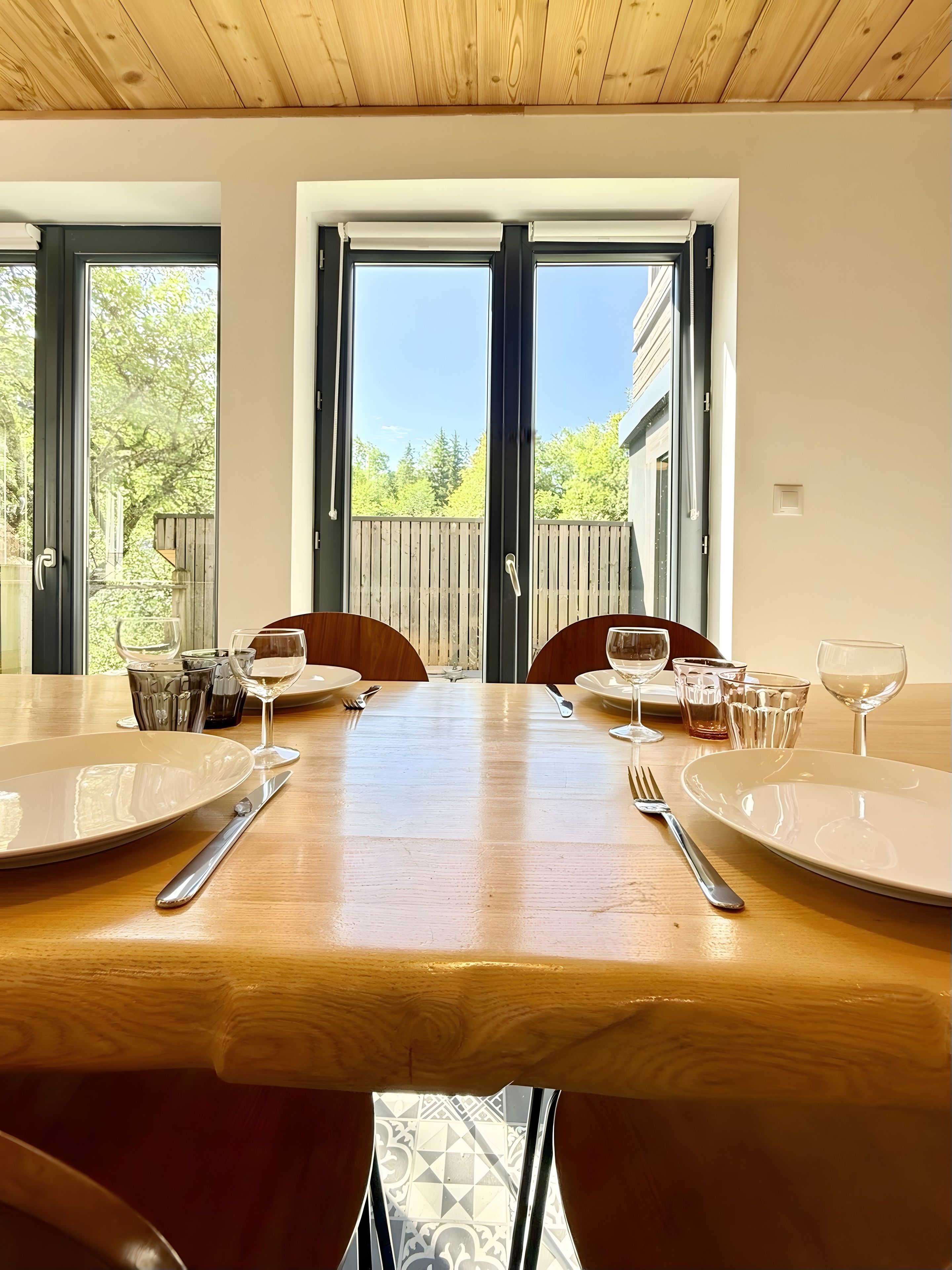 A dining table set for two with plates, cutlery, and glasses, positioned in a room with wooden ceiling and large windows.