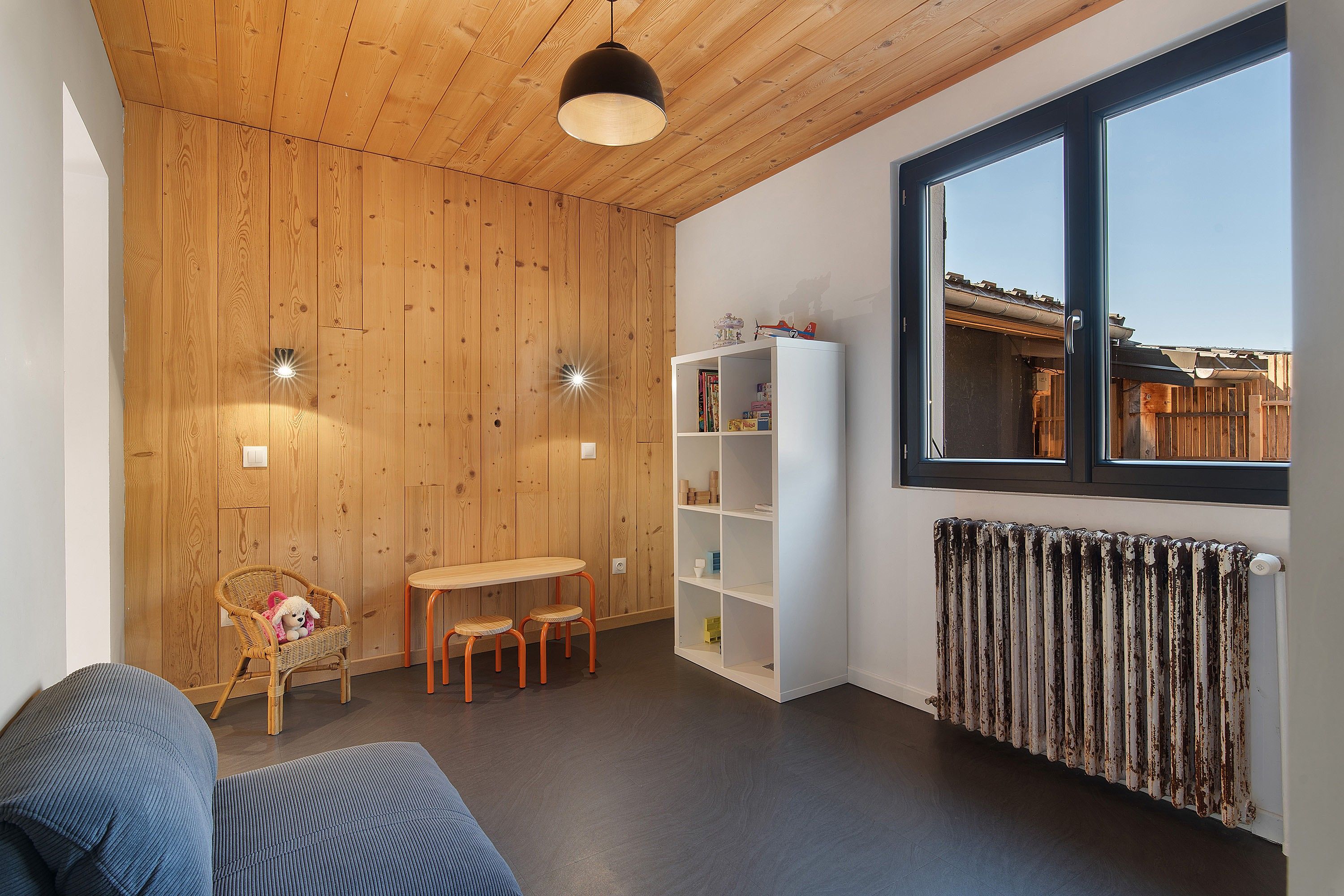 Une pièce aux murs en bois, une bibliothèque blanche, une table avec des chaises, un fauteuil en osier avec un jouet en peluche, et une fenêtre donnant sur un immeuble.