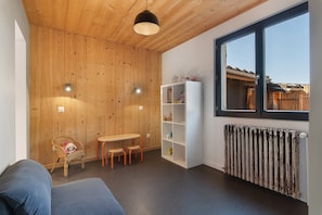 A room with wooden walls, a white bookshelf, a table with chairs, a wicker chair with a stuffed toy, and a window with a view of a building.