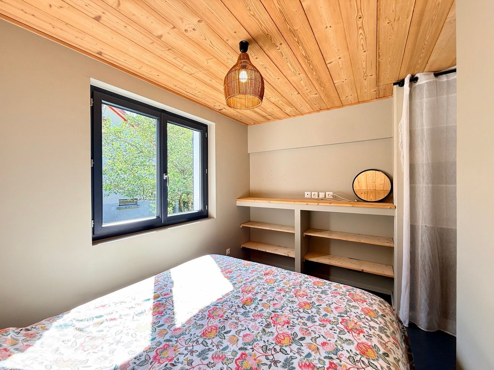 A bedroom with a bed, a window, a desk with a mirror, and a wooden ceiling.