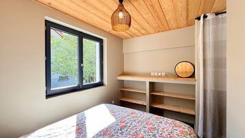A bedroom with a bed, a window, a desk with a mirror, and a wooden ceiling.