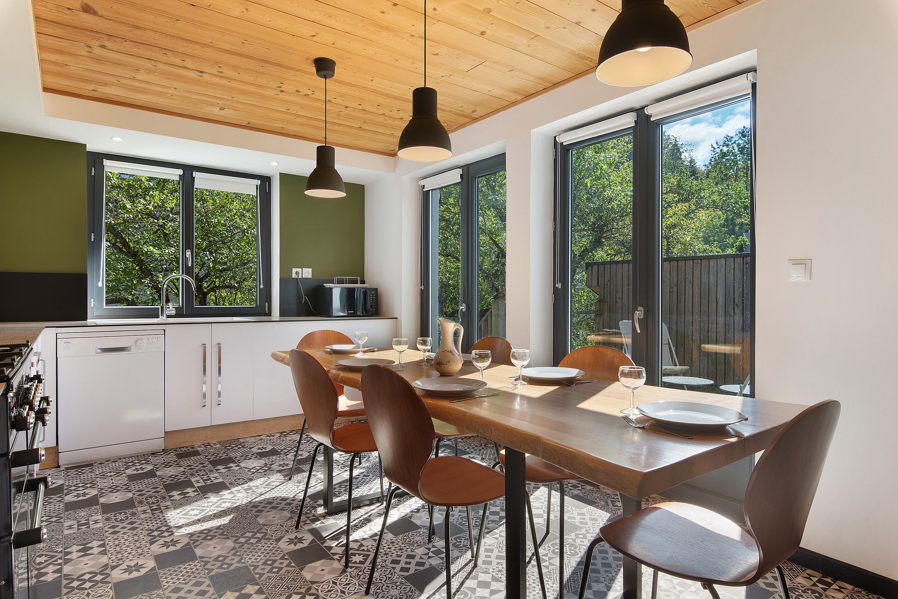 Eine moderne Küche mit weißen Schränken, ein Essbereich mit Holztisch und -stühlen sowie große Fenster mit Blick ins Grüne.