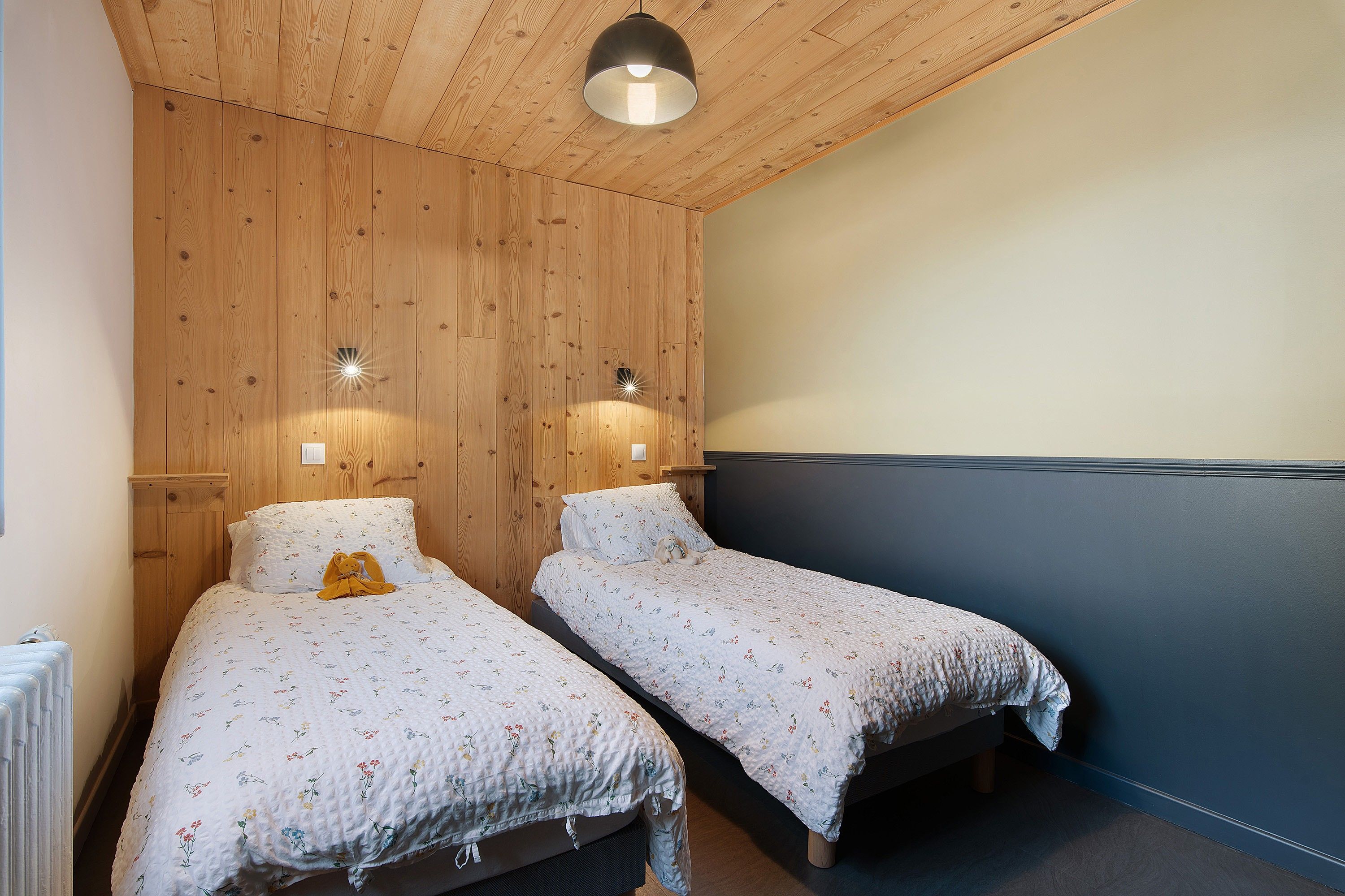 Une chambre avec deux lits simples, des murs recouverts de panneaux de bois, un luminaire au plafond et un radiateur.