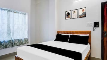 A hotel room with a bed, a window with a floral curtain, a wall-mounted power outlet, and a door with a wooden frame.