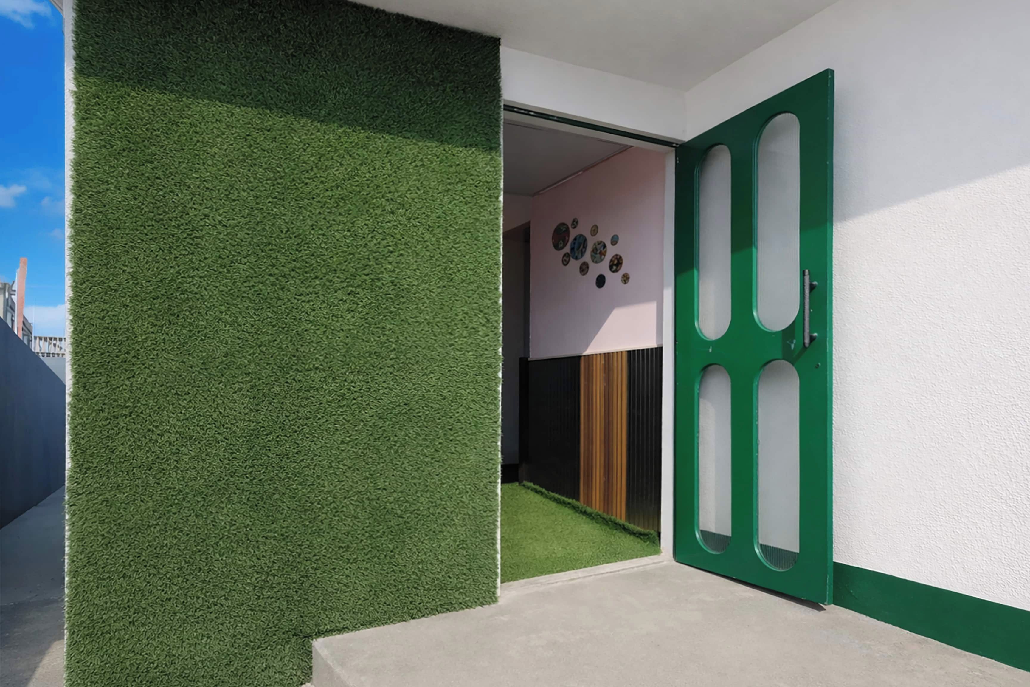 Entrada de un edificio moderno con un muro de césped artificial verde, una puerta verde con paneles recortados y una habitación con suelo de madera y pared decorada con objetos circulares.