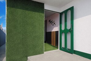 A modern building entrance with a green artificial grass wall, a green door with cut-out panels, and a room with a wooden floor and a wall decorated with circular objects. - Collection O Jalukbari Bus Stand (Guwahati)