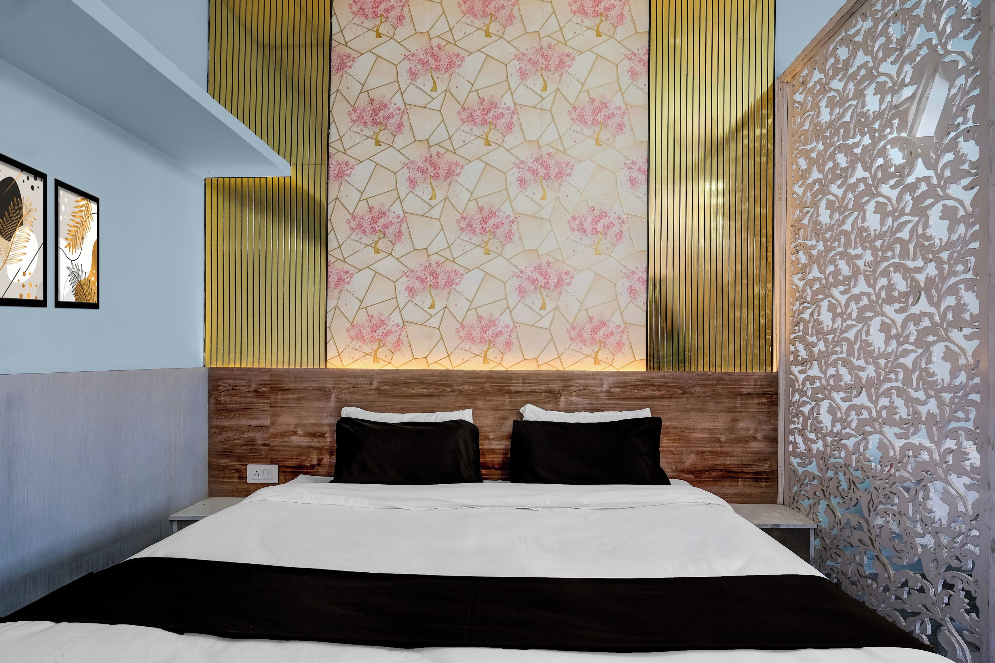 A hotel room with a bed, two pillows, a wooden headboard, a decorative wall with a geometric pattern, and a window with vertical blinds.