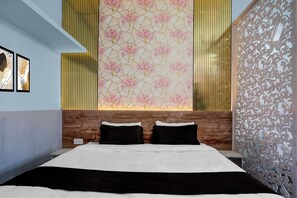 A hotel room with a bed, two pillows, a wooden headboard, a decorative wall with a geometric pattern, and a window with vertical blinds.