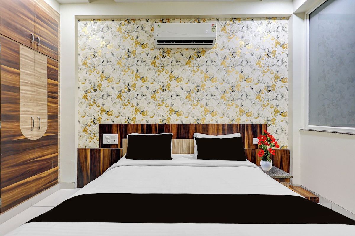 Un dormitorio con cama, cabecera de madera, papel tapiz floral, unidad de aire acondicionado y mesita de noche con un florero.