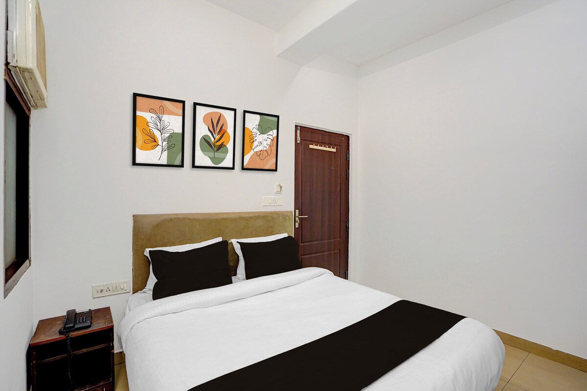 A hotel room with a bed, a wooden nightstand, a telephone, and a door. There are three framed artworks on the wall.