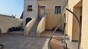 A residential building with a staircase leading to a door, a small balcony with a flower planter, and a metal chair.