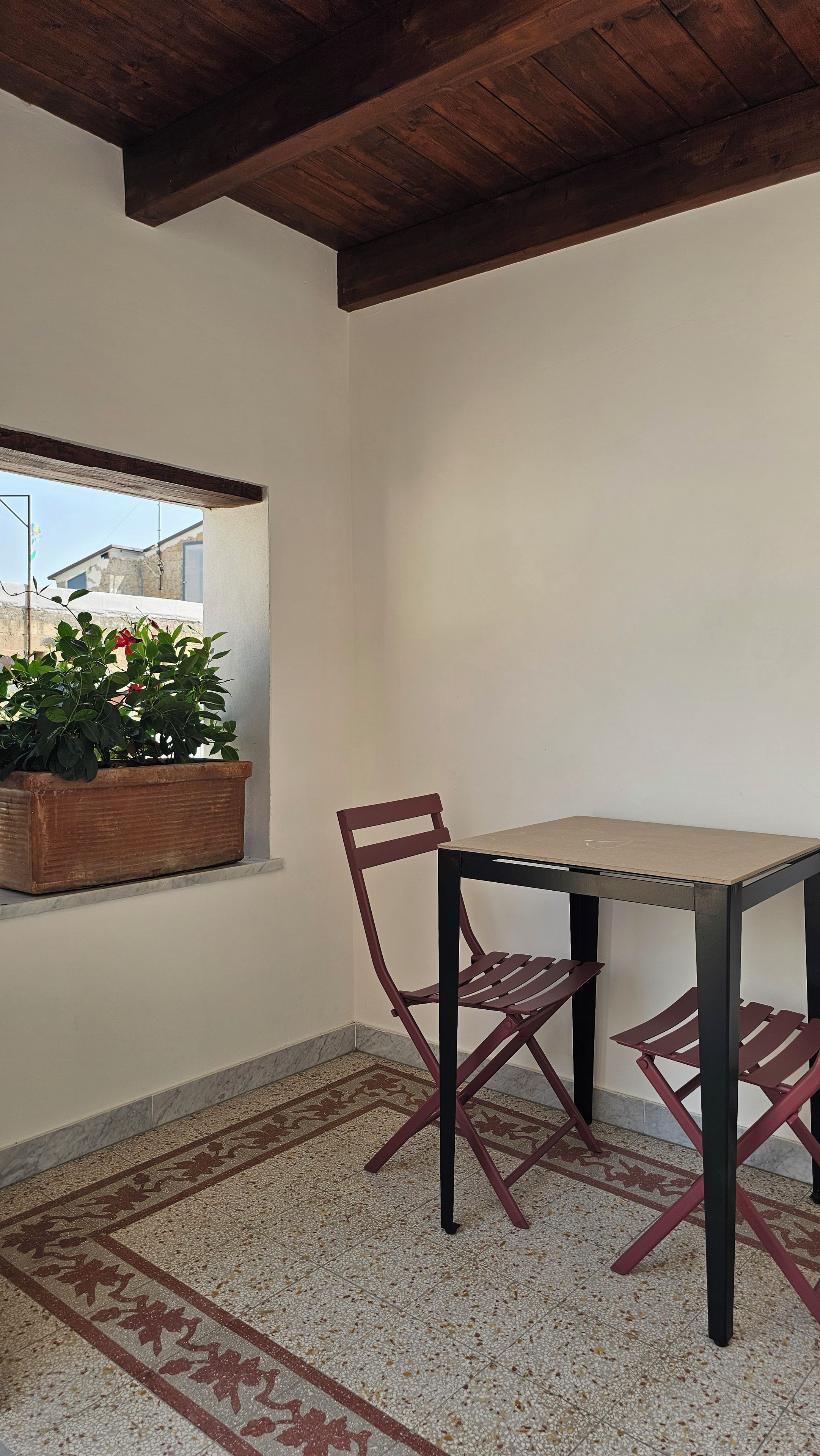Habitación de esquina con una mesa, dos sillas, una planta en maceta y ventana con vistas a edificios.