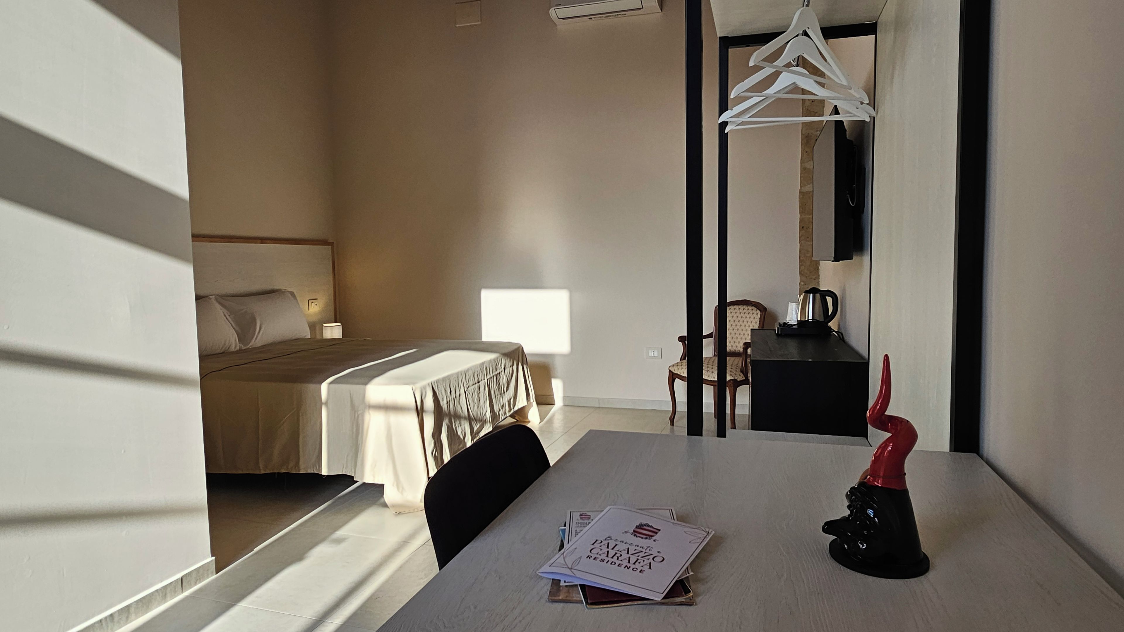 Hotelværelse med seng, bord med menu, stol, knagerække og en rød dekorativ genstand.