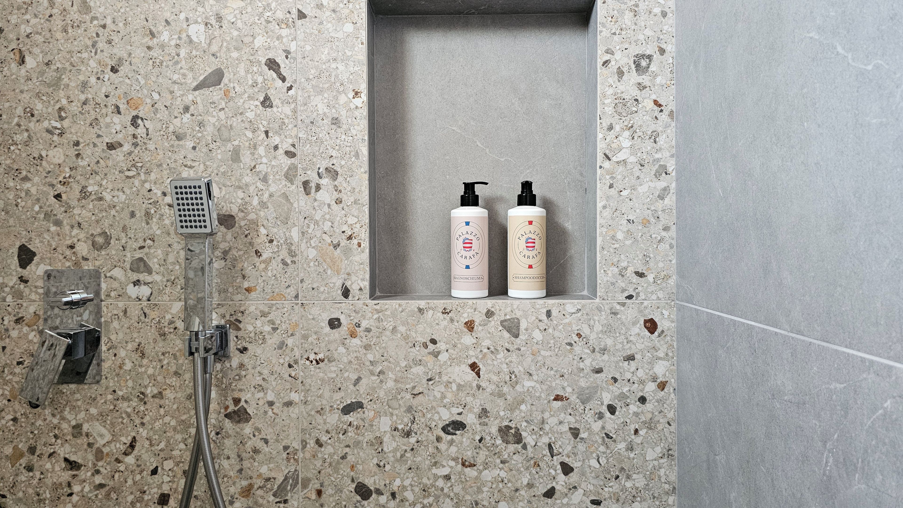 Un baño con ducha, un estante empotrado con dos botellas de artículos de aseo, y un cabezal de ducha de mano.