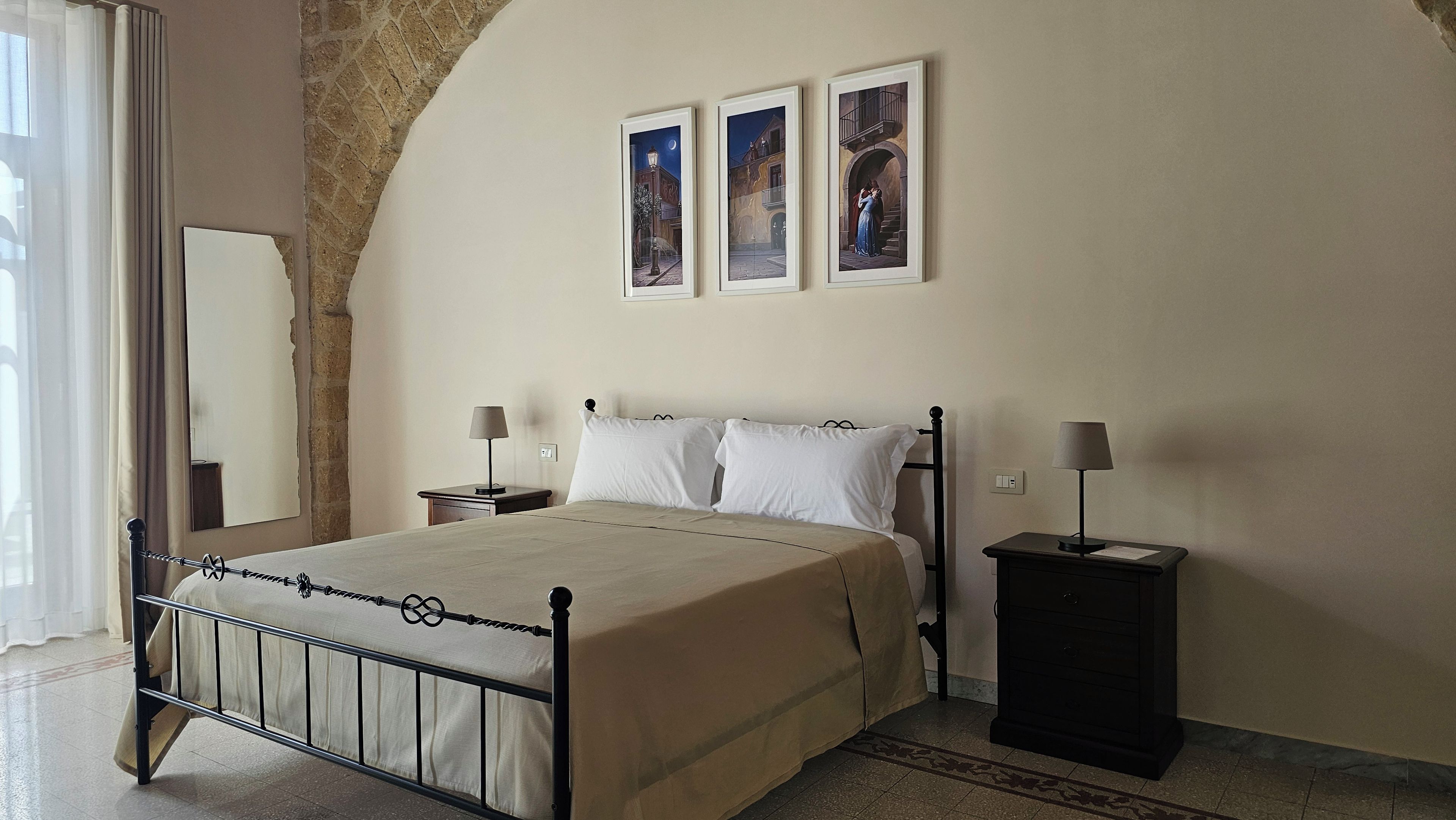 A bedroom with a bed, two bedside tables, a nightstand, a mirror, and three framed pictures on the wall.