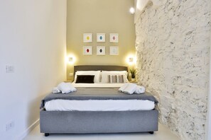 Uma cama bem arrumada com colchão cinza e roupa de cama branca, cabeceira com almofadas decorativas e parede com revestimento de pedra texturizado.