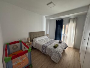 Ein Schlafzimmer mit einem Bett, einem Nachttisch, einer Lampe, einem Fenster mit Vorhängen und einem Spielbereich für Kinder.