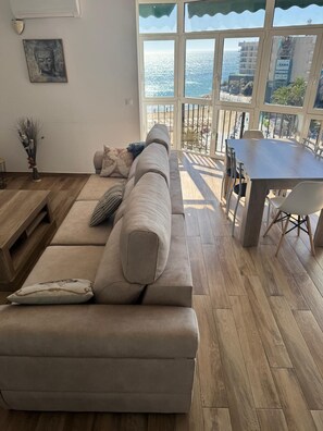Ein Wohnzimmer mit einem beigen Sofa, einem Holztisch und einem Esstisch mit Stühlen. Ein großes Fenster bietet Blick auf das Meer und Gebäude.