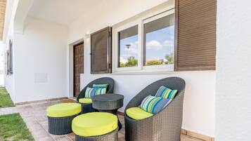 A patio area with wicker furniture and cushions, a window with blinds, and a white exterior wall.