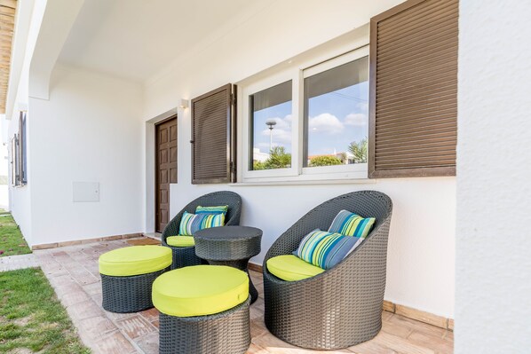 A patio area with wicker furniture and cushions, a window with blinds, and a white exterior wall. - Villa 'Grandma's Beach' with Sea View, Wi-Fi and Air Conditioning (Albufeira)
