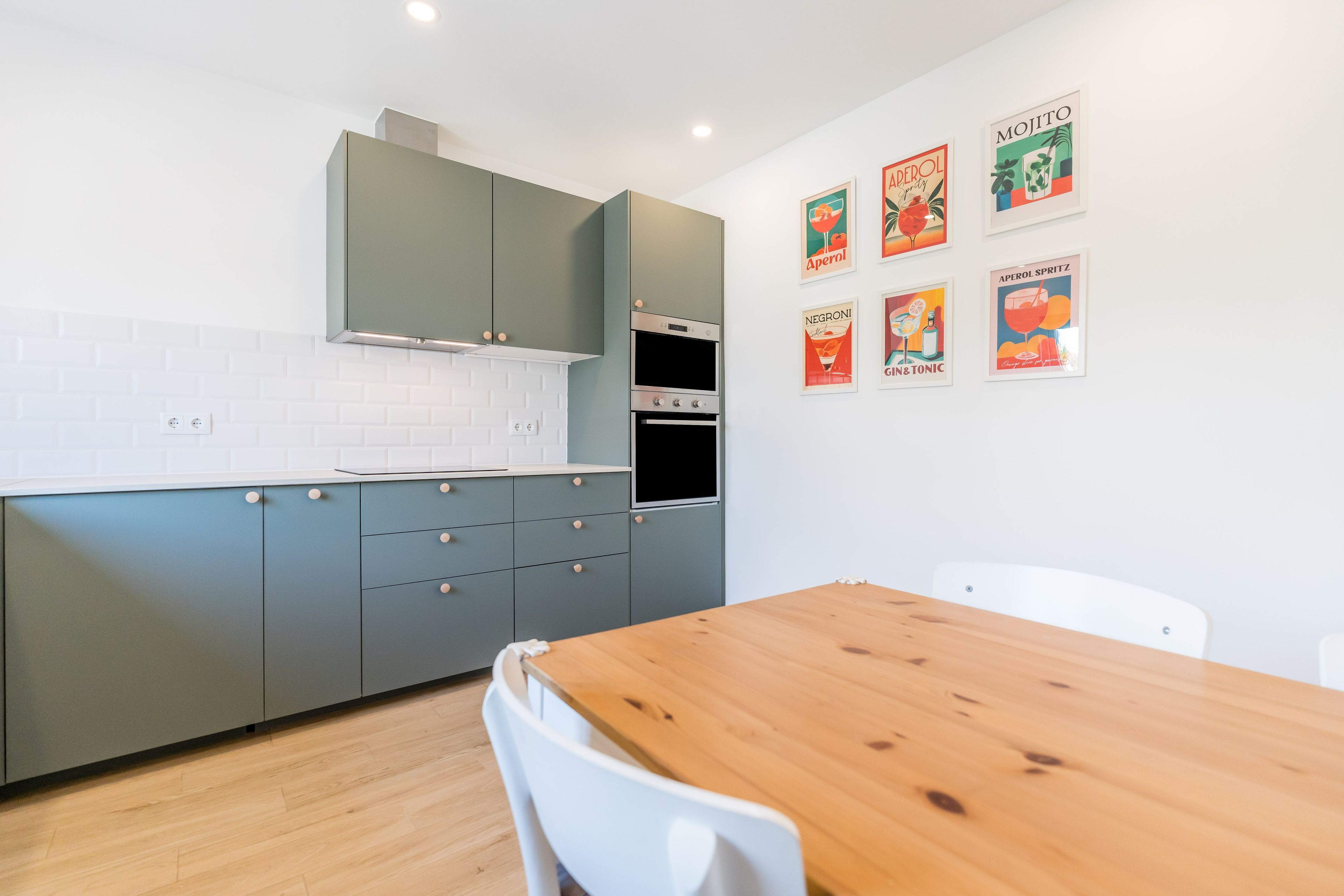 A modern kitchen with grey cabinets, a wooden dining table, and a white chair. There are six framed pictures on the wall.