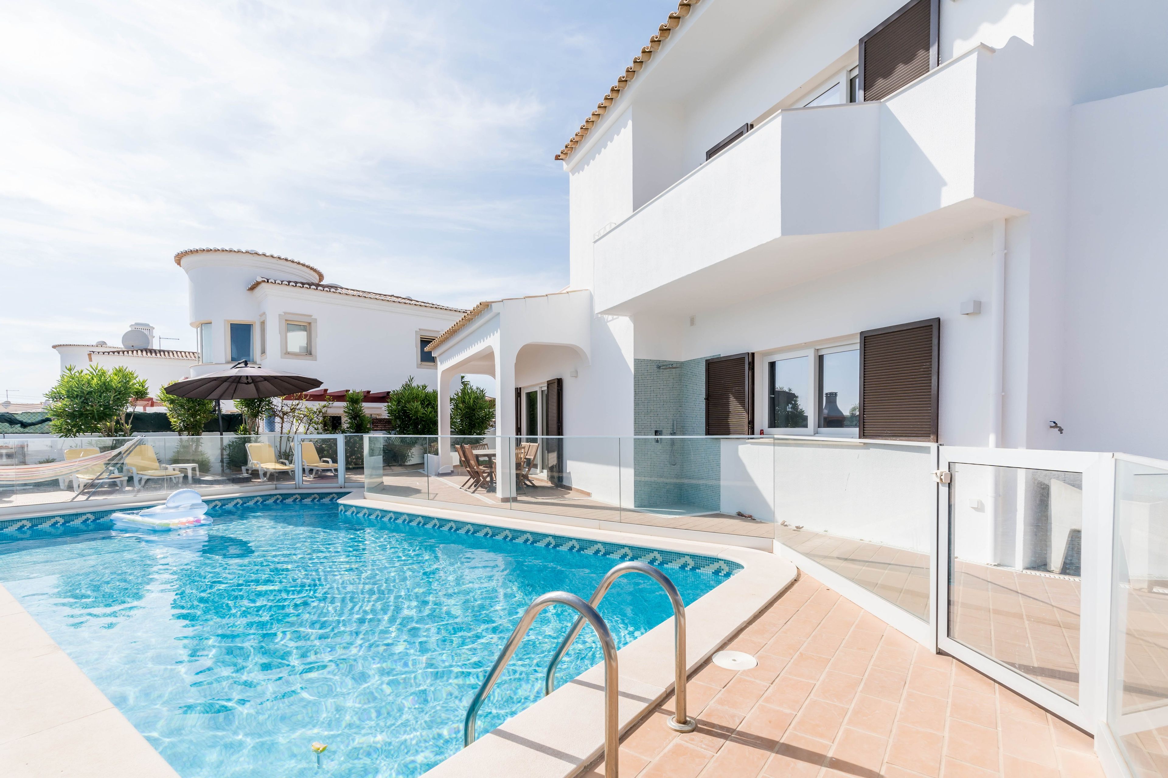 A swimming pool with a glass balustrade, a sun umbrella, and lounge chairs.