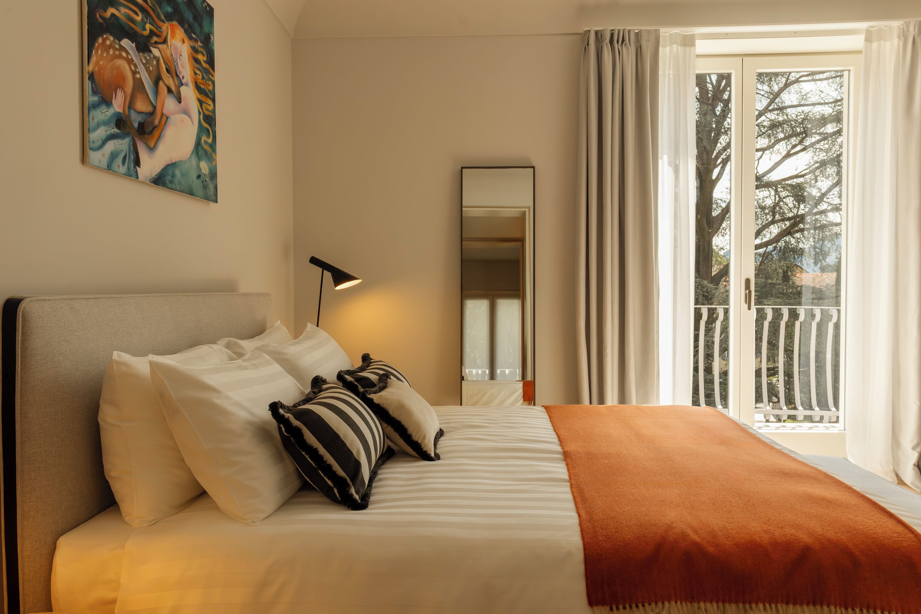 Hotellihuone, jossa on sänky, tyynyjä, peitto, lamppu, peili ja näkymä ulos.