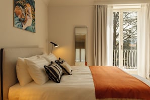 A hotel room with a bed, pillows, a blanket, a lamp, a mirror, and a view of the outdoors.