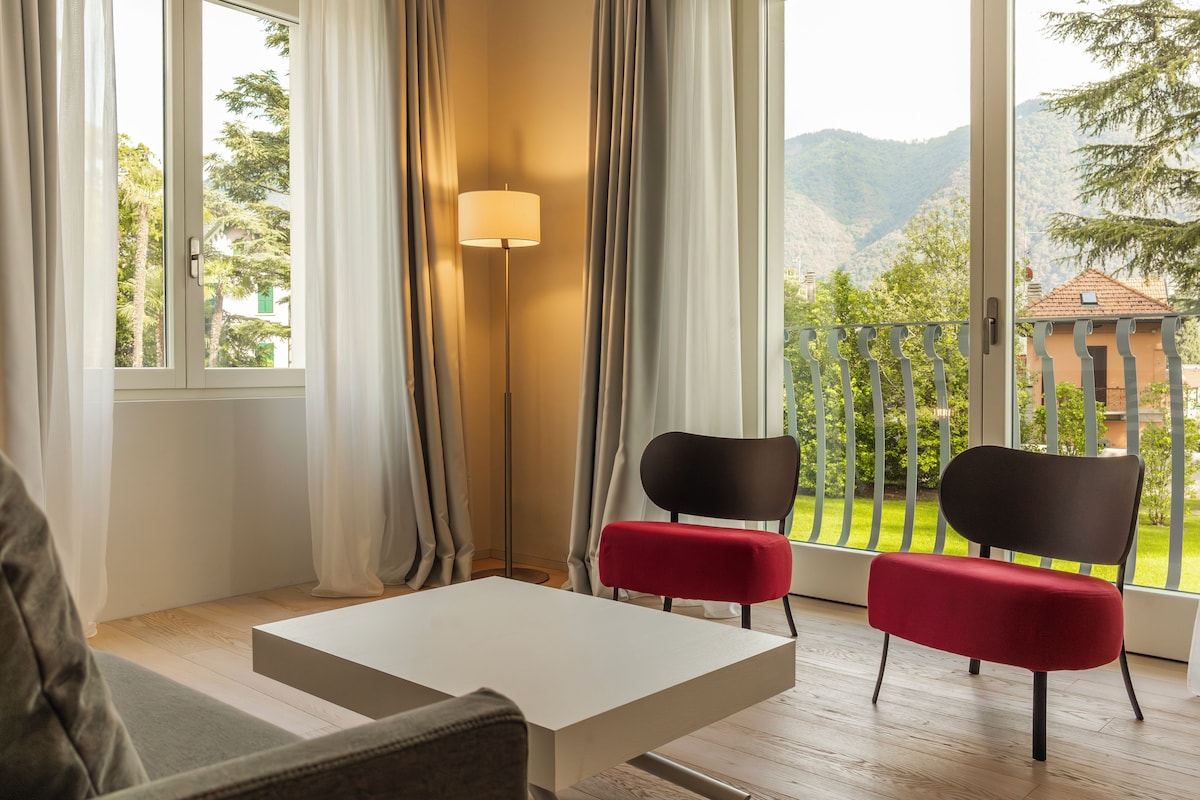 A room with a view of a mountainous landscape, a floor lamp, two chairs, and a table.