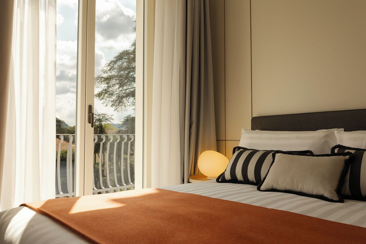 A hotel room with a bed, a nightstand, a lamp, and a view of the outdoors through a window with curtains.