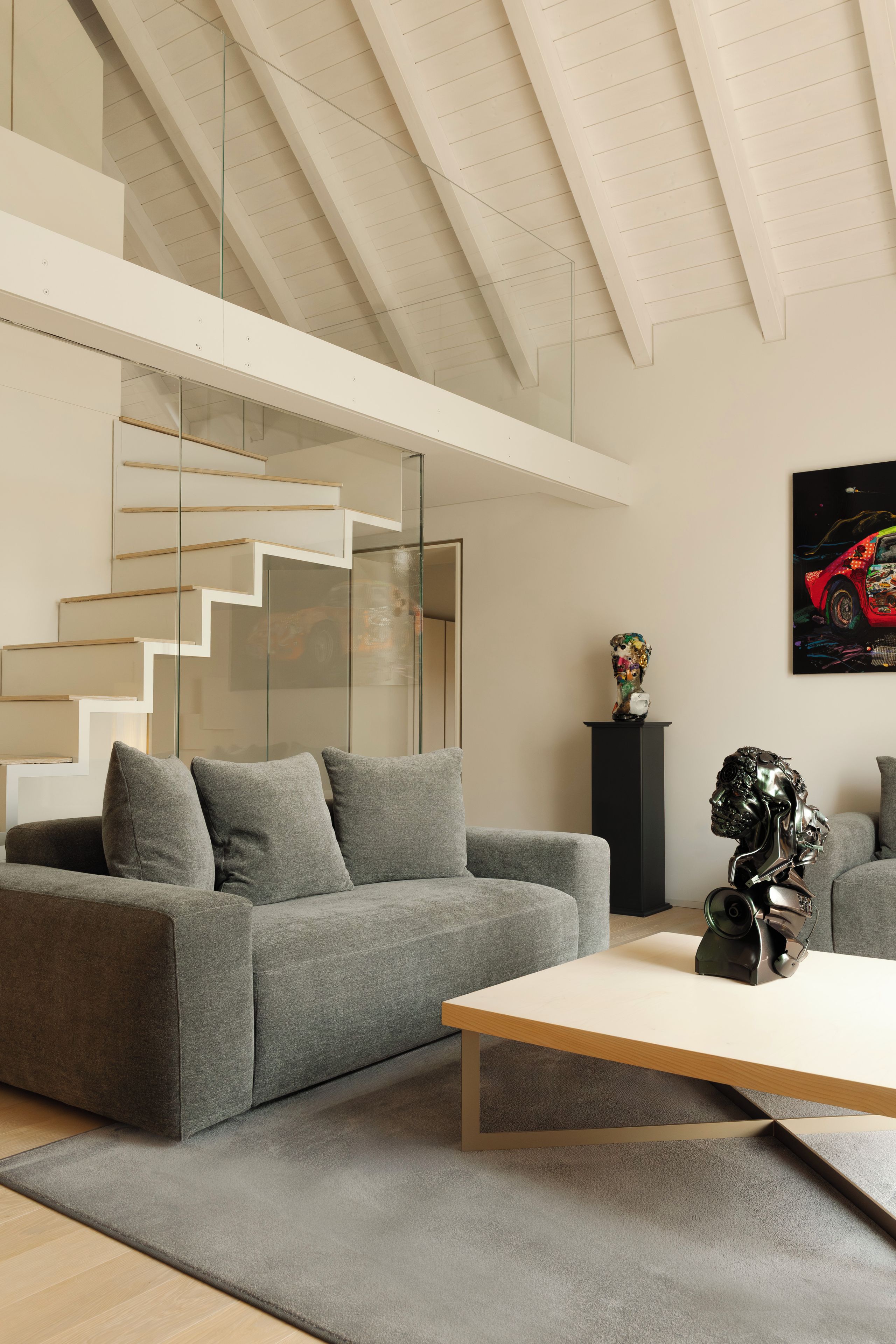 Una sala moderna con un sofá gris, una mesa de centro de madera y una escalera con barandillas de cristal.