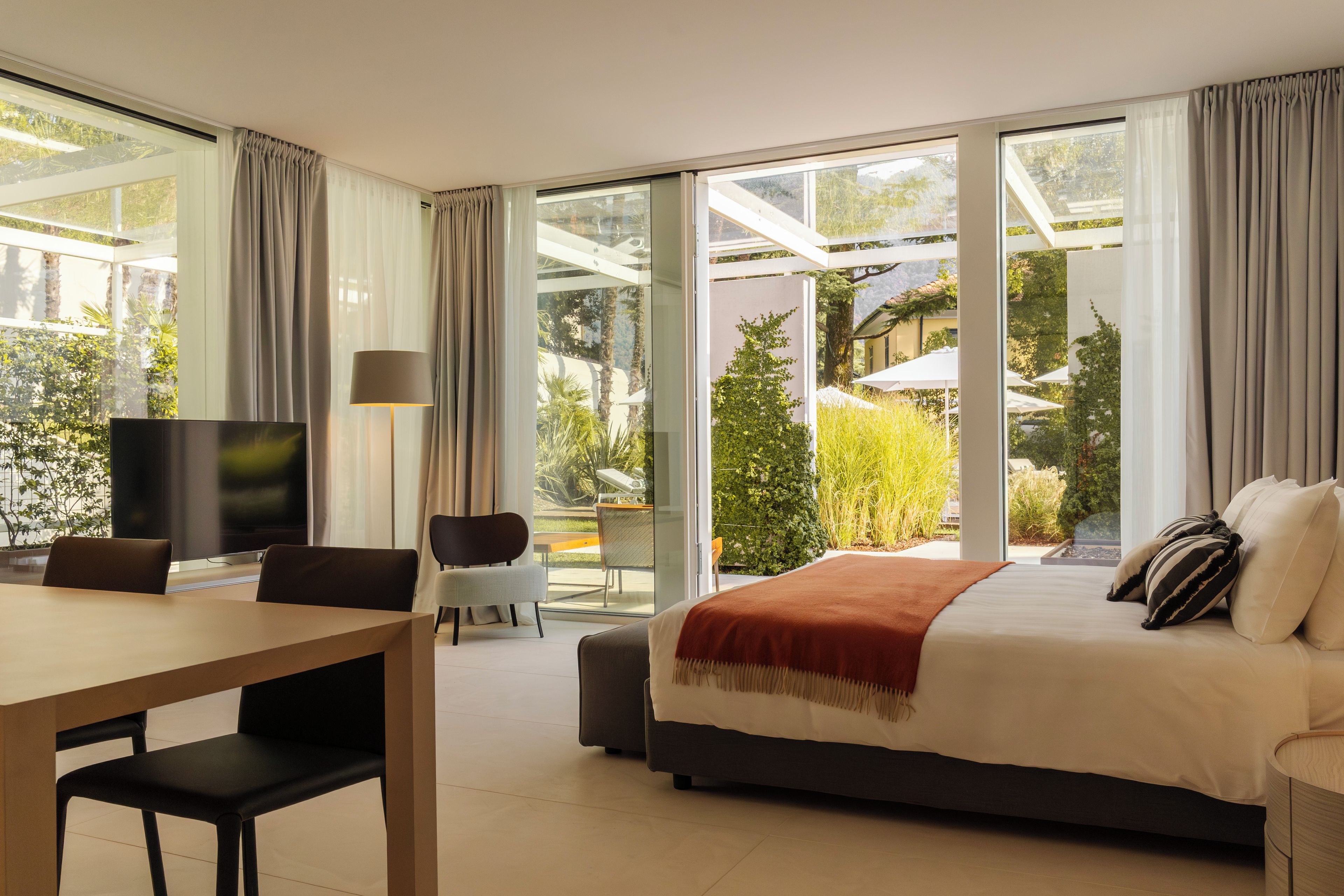 A modern hotel room with a large bed, a desk with a chair, a television, and a view of a garden through large windows.