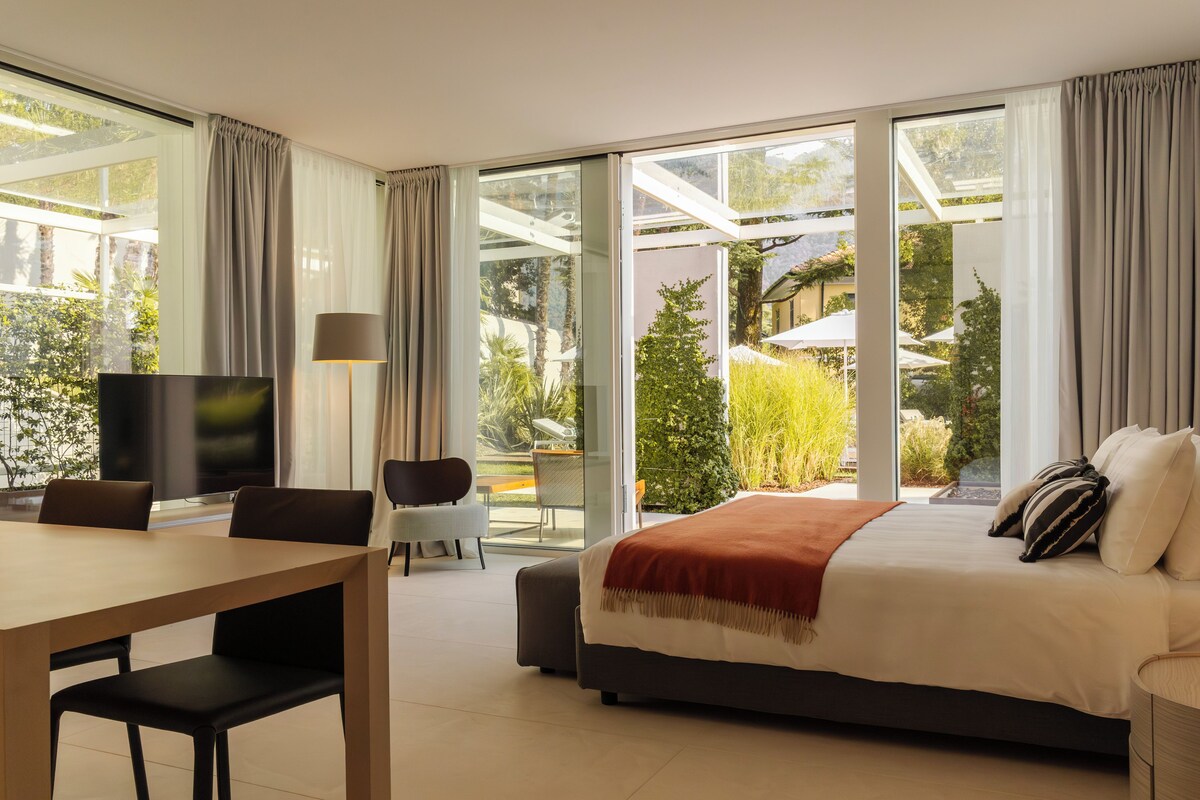 A modern hotel room with a large bed, a desk with a chair, a television, and a view of a garden through large windows.