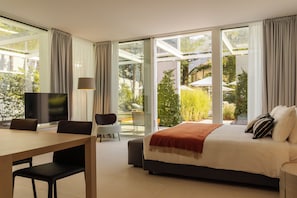 A modern hotel room with a large bed, a desk with a chair, a television, and a view of a garden through large windows.