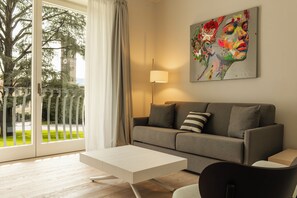 A modern living room with a grey sofa, a white coffee table, and a lamp. There is a painting on the wall and a view of a tree and a building outside.