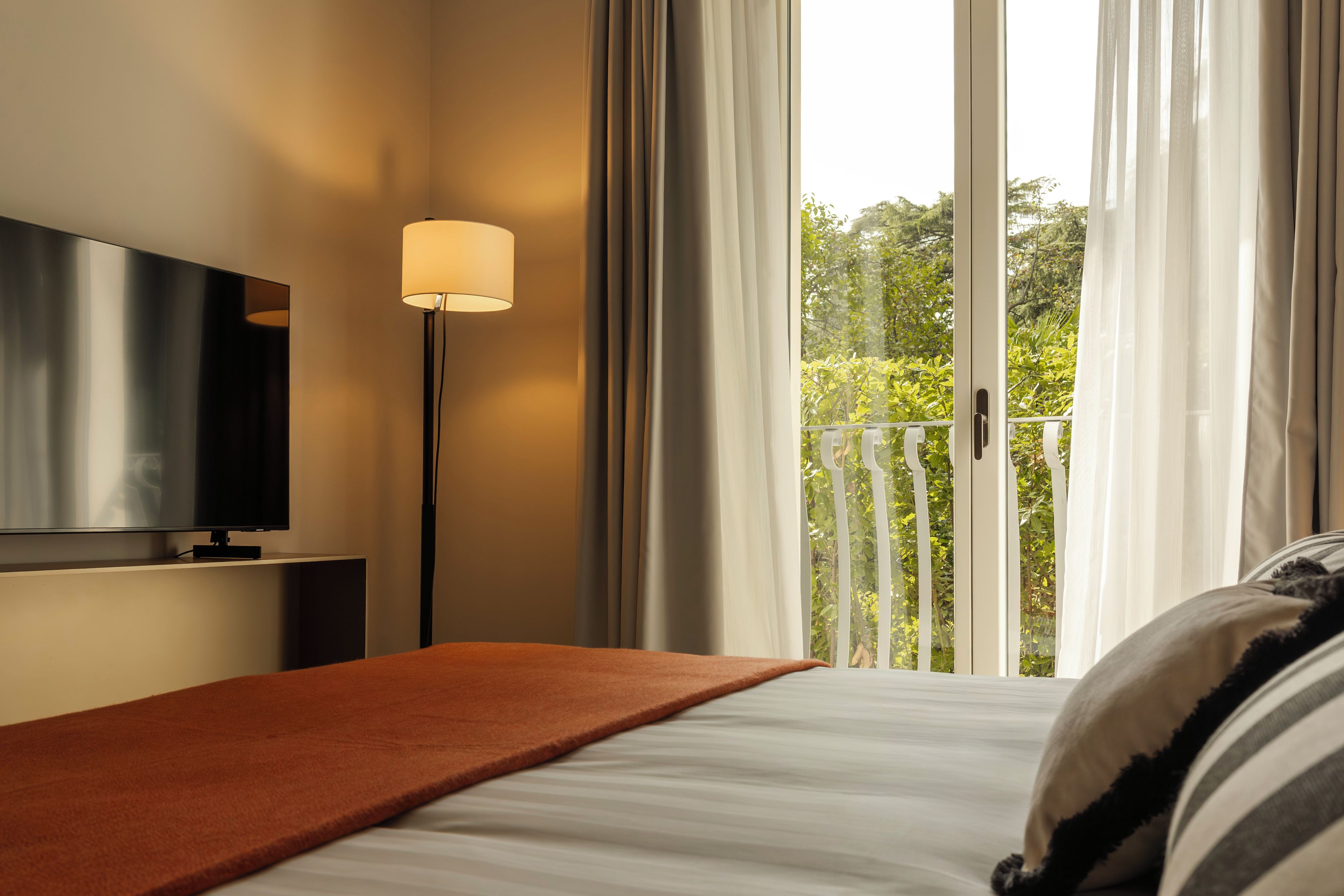A hotel room with a bed, a TV mounted on the wall, a floor lamp, and a balcony with a view of greenery.