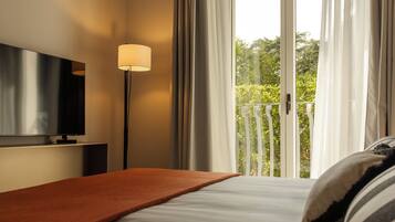 A hotel room with a bed, a TV mounted on the wall, a floor lamp, and a balcony with a view of greenery.