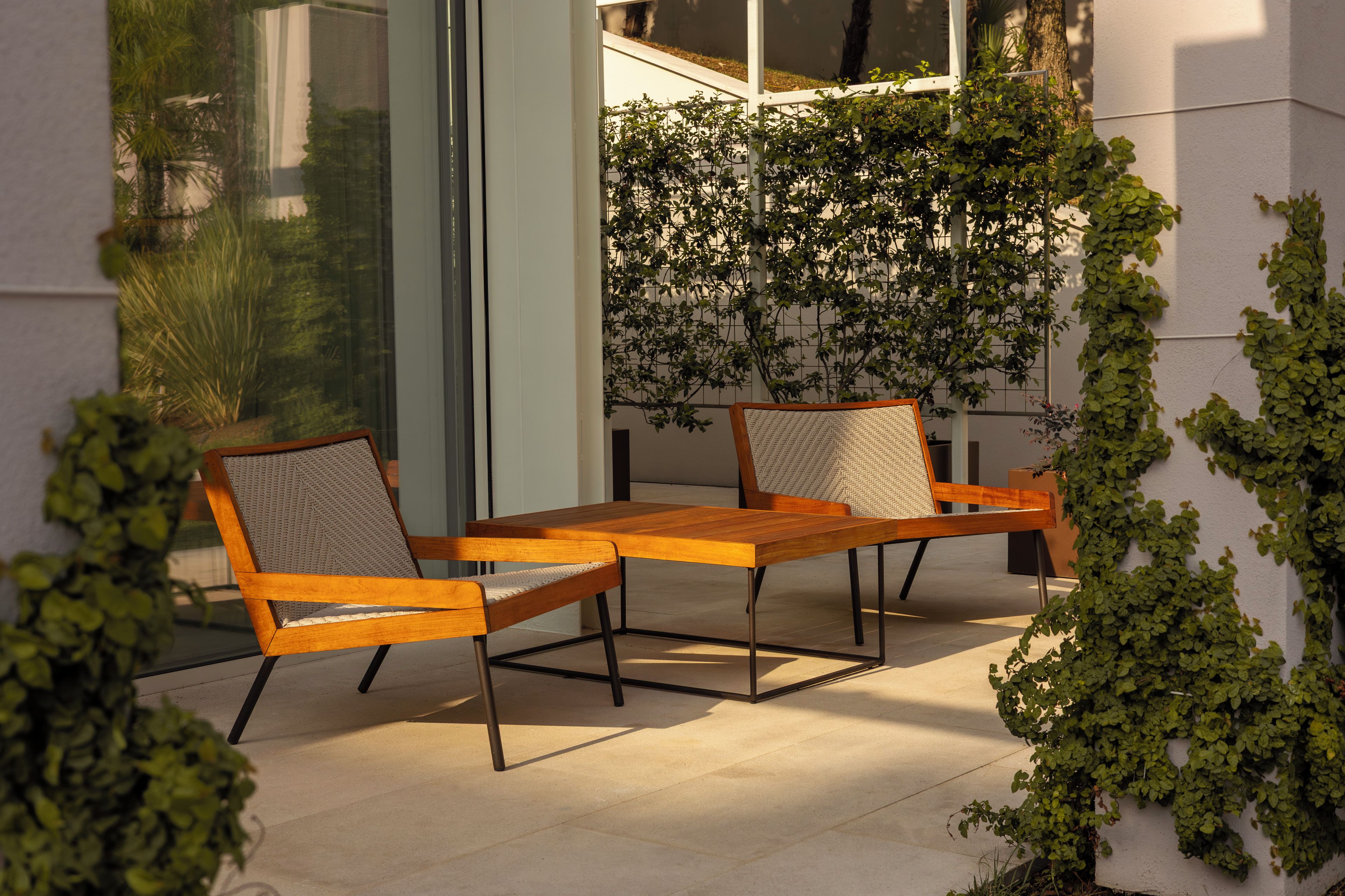 An outdoor seating area with two wooden tables and chairs, surrounded by greenery and a glass wall.