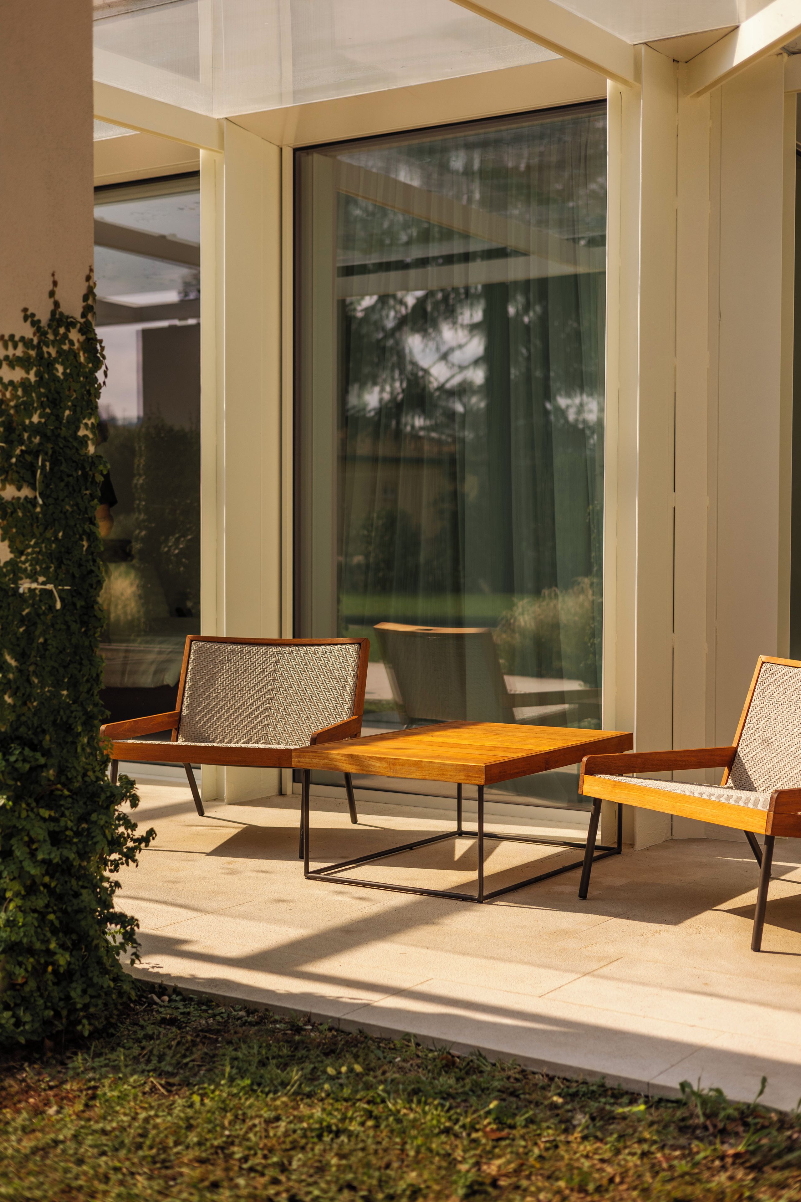 Un área de estar moderna al aire libre con dos sillas y una mesa, rodeada por un cerramiento de vidrio y vistas a árboles.