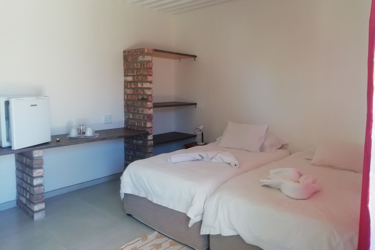 Un dormitorio con una cama, un escritorio pequeño con refrigerador, un pilar de ladrillo y una mesita de noche con lámpara.