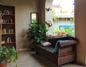 Un rincón acogedor para leer, con un sofá de mimbre, una estantería llena de libros y una planta grande en maceta.