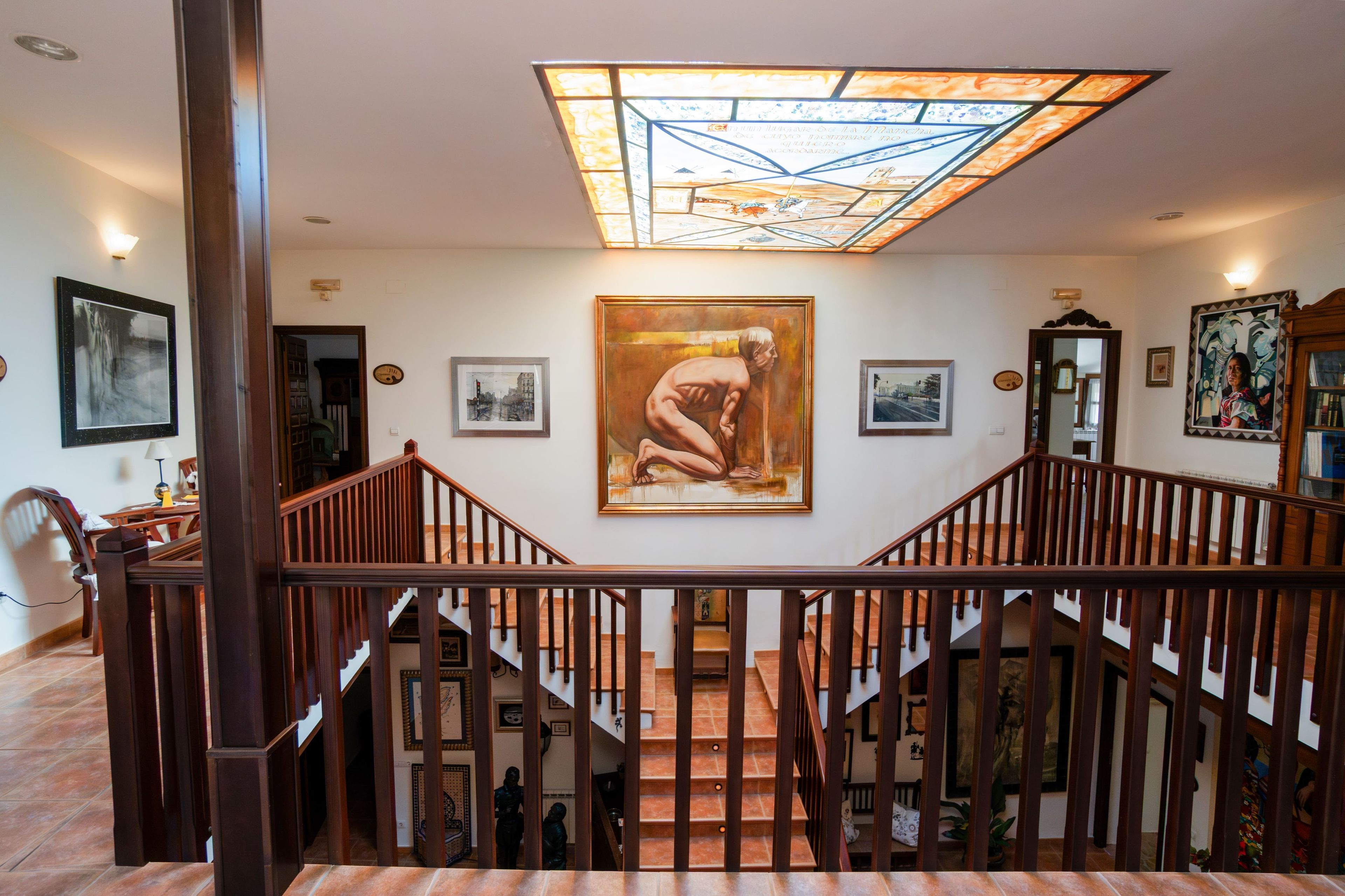 Una escalera con barandillas de madera que conduce a un piso superior, un cuadro grande en la pared y un techo de vitrales.