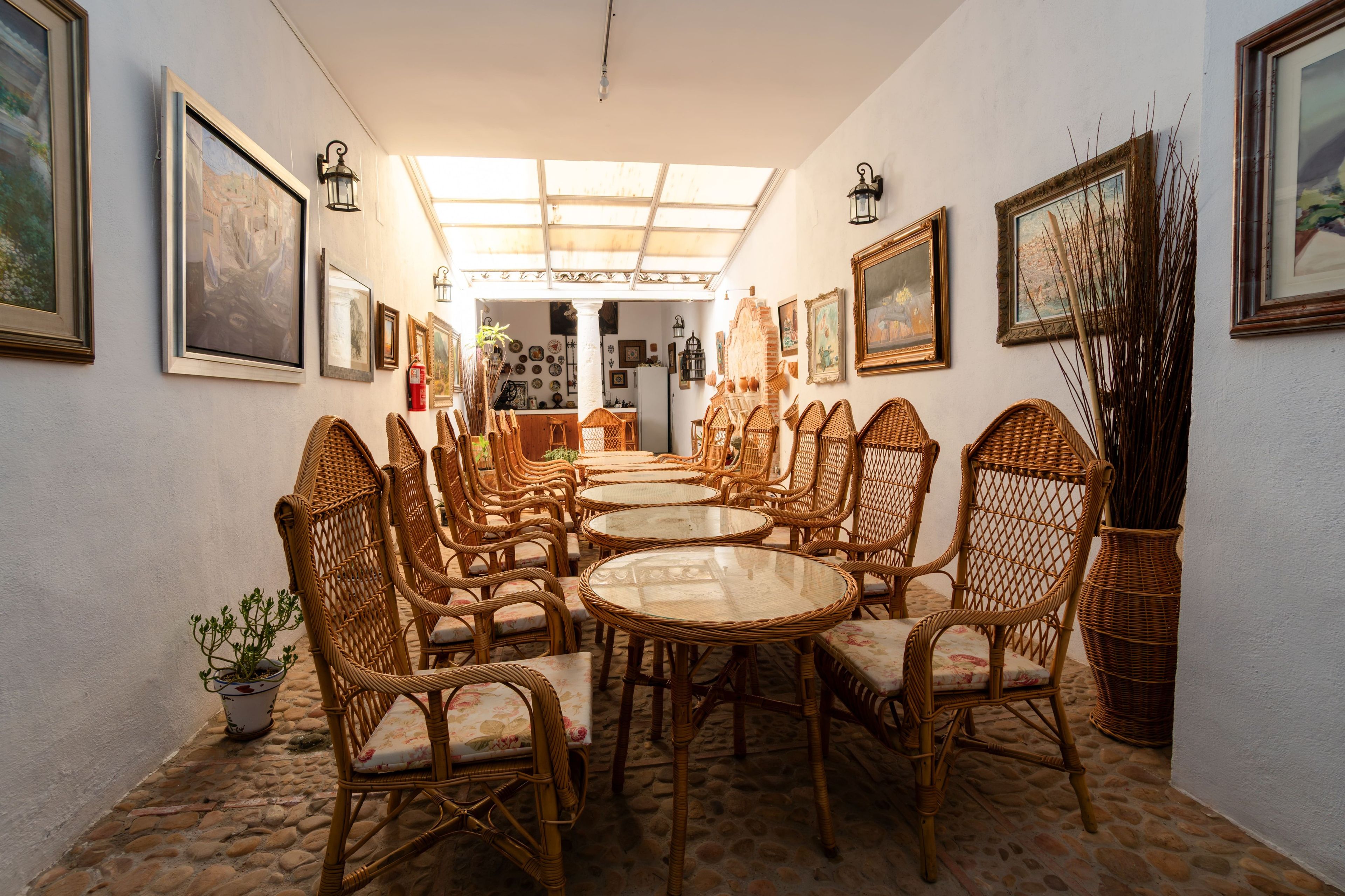 Un comedor con sillas de mimbre y mesas redondas, obras de arte en las paredes y un tragaluz.
