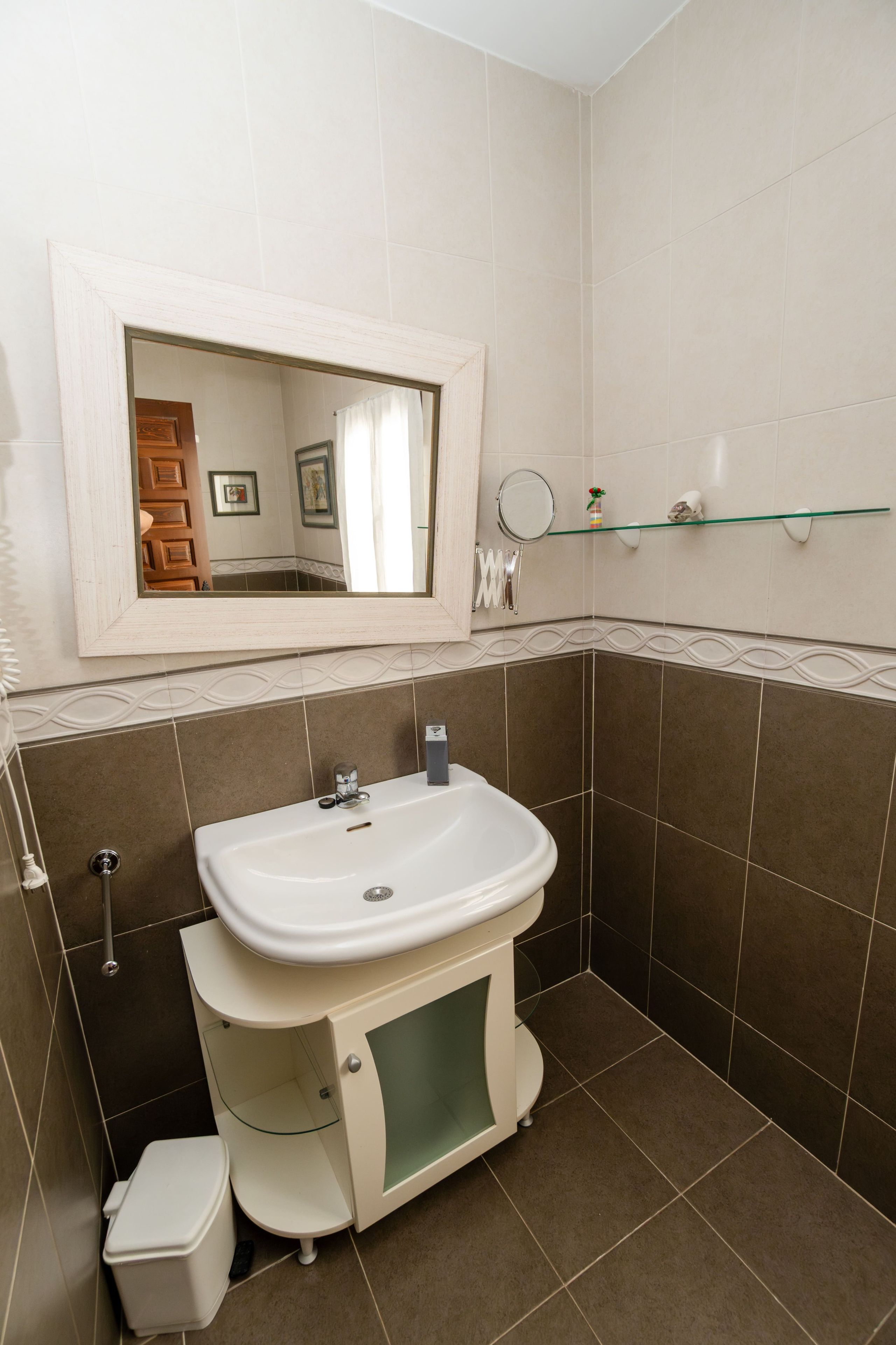 Un bagno con un lavandino bianco, uno specchio sopra e uno scaffale in vetro con articoli da toilette.