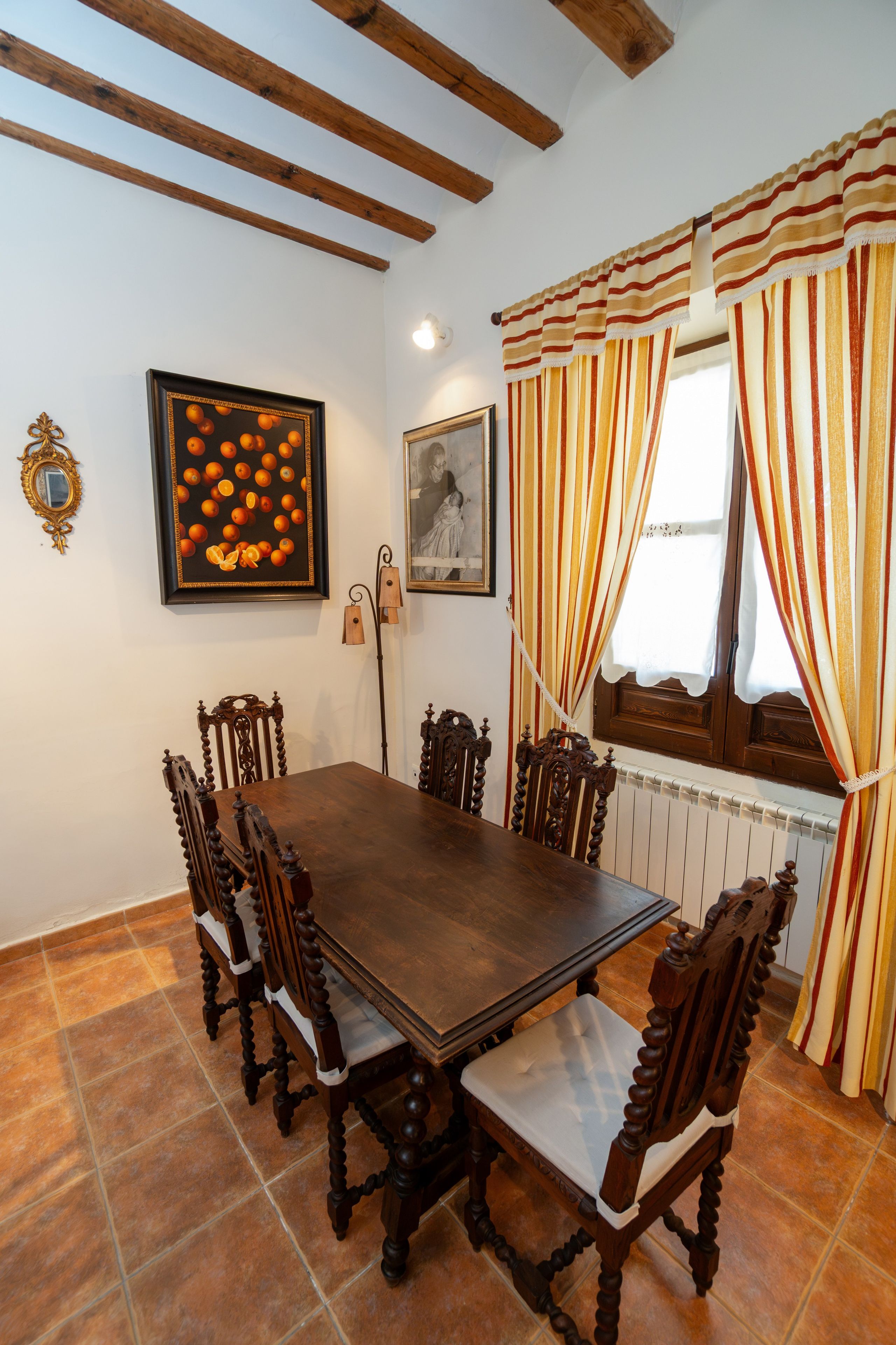 Un comedor con una mesa y sillas de madera, un cuadro enmarcado con naranjas y una ventana con cortinas a rayas.